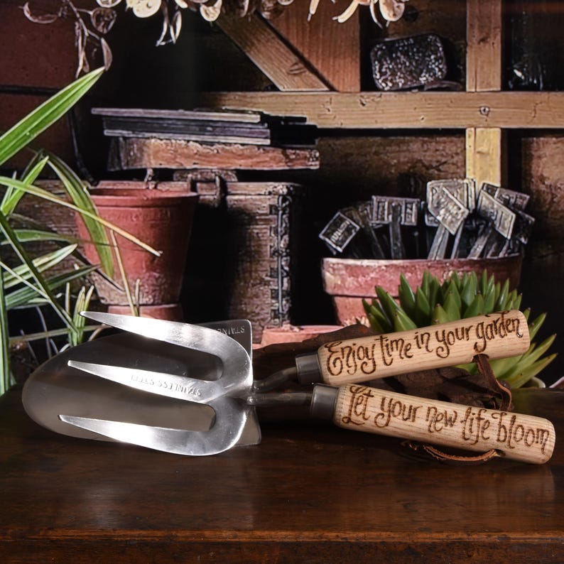 May include: A stainless steel garden hand fork and two wooden-handled garden tools. The wooden handles have the phrases "Enjoy time in your garden" and "Let your new life bloom" etched on them. The tools are on a dark wooden surface.