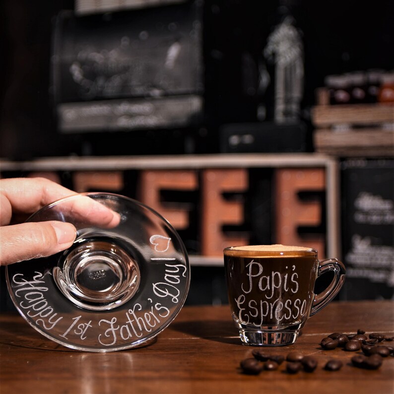 May include: A clear glass espresso cup and matching saucer set. The cup features the white etched text Papi's Espresso, while the saucer is etched with the text Happy 1st Father's Day and a small heart icon. The set is made of transparent glass.
