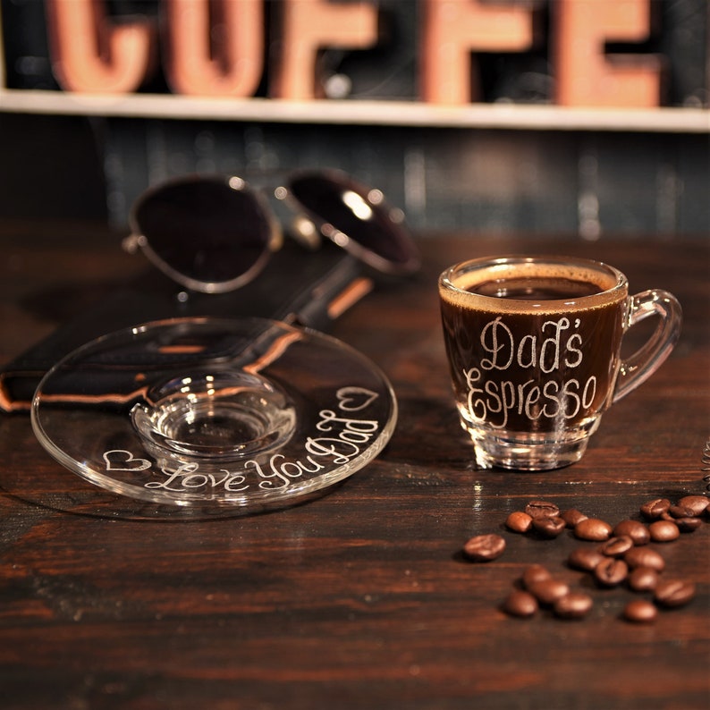 May include: A clear glass espresso cup and matching saucer set. The cup features the white etched text "Dad's Espresso" and the saucer is etched with the text "Love You Dad" accompanied by two small heart icons, filled with dark coffee.