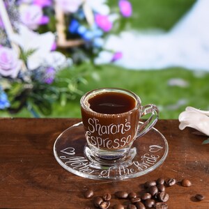 May include: A clear glass espresso cup and matching saucer filled with dark coffee. The cup features the white etched text Sharon's Espresso, while the saucer displays the white etched text Mother of the Bride with two small heart icons.