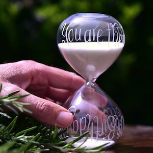 May include: A clear glass hourglass with white sand. The top of the hourglass is engraved with the words "You are the" and the bottom is engraved with the words "Happiest Girl".