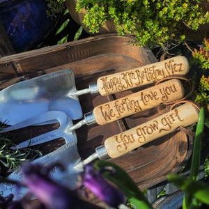 May include: Three stainless steel garden tools with wooden handles. The handles are engraved with the phrases "Happy 30th Birthday Mez", "We forking love you", and "You grow girl". The tools are arranged in a wooden tray.