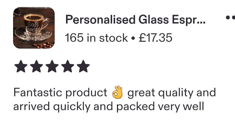 May include: A clear glass espresso cup and matching saucer set featuring the white text "Dad's Espresso" printed on the side of the cup. The cup is filled with dark brown coffee and is positioned next to several scattered roasted coffee beans.