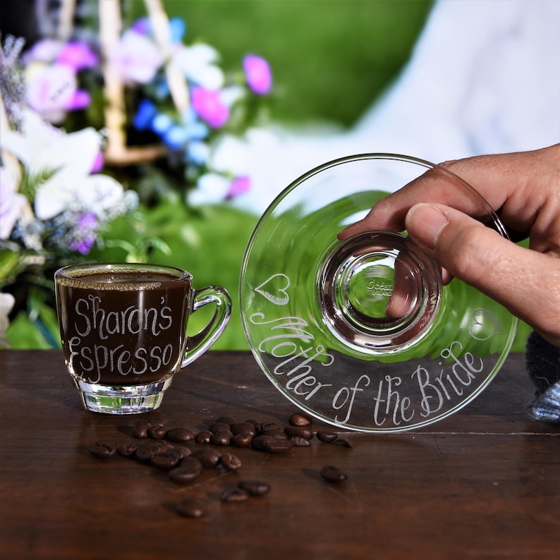 May include: A clear glass espresso cup and matching saucer. The cup is filled with dark coffee and engraved with the text "Sharon's Espresso". The saucer is engraved with a heart icon and the text "Mother of the Bride" in cursive script.