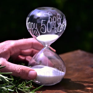 May include: A clear glass hourglass with white sand inside. The hourglass has the words "Happy 50th Birthday" etched on the glass. The hourglass is held by a hand, with rosemary sprigs in the foreground.