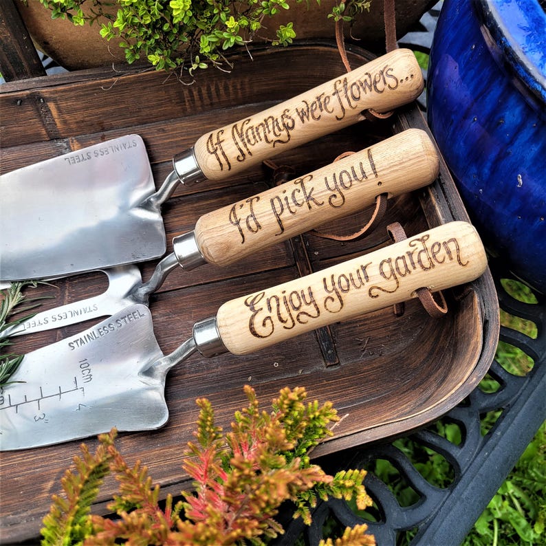 May include: Three stainless steel garden trowels with wooden handles. The handles are engraved with the phrases "If Nannys were flowers...", "I'd pick you!", and "Enjoy your garden". The trowels are displayed on a wooden tray.