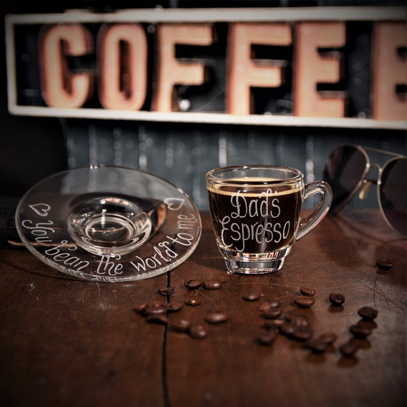 May include: A clear glass espresso cup and matching saucer set. The cup features the white etched text "Dad's Espresso" and the saucer is etched with "You bean the world to me" along with small heart icons. The cup is filled with dark coffee.