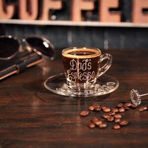 May include: A clear glass espresso cup and matching saucer filled with dark coffee. The cup features the white text Dad's Espresso, while the saucer is etched with the phrase Love You Dad. Scattered roasted coffee beans surround the set.