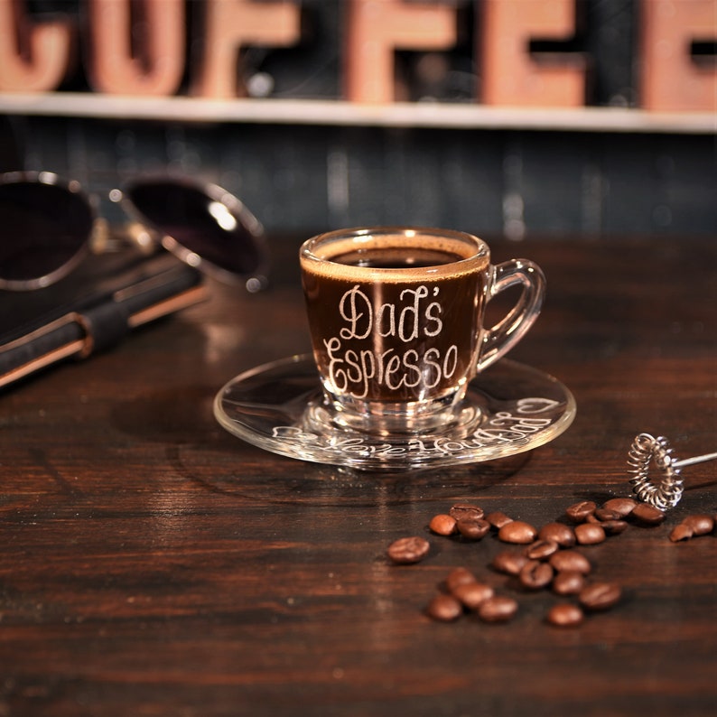 May include: A clear glass espresso cup and matching saucer filled with dark coffee. The cup features the white text Dad's Espresso, while the saucer is etched with the phrase Love You Dad. Scattered roasted coffee beans surround the set.