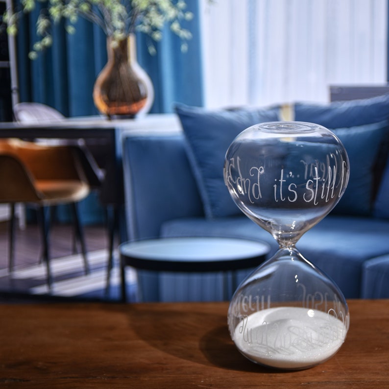 May include: A clear glass hourglass with white sand inside. The hourglass has etched text that reads "...and it's still..." and "...time..." on the top and bottom bulbs. The hourglass is on a wooden surface, with a blue background.