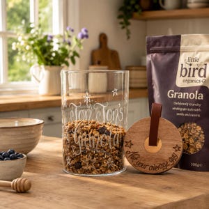 May include: A clear glass canister filled with granola, featuring white etched stars and the text "Jon Jon's Granola". Beside it sits a round wooden lid with a brown leather strap and the etched text "Not your average guy".