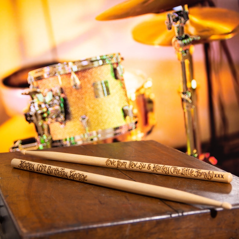 A pair of wooden drum sticks rest in front of a yellow and brass drumkit.  The drumsticks have been hand engraved in a calligraphy font with the words Happy 18th Birthday George on one stick, love from Trev, Sue and Harley on the other