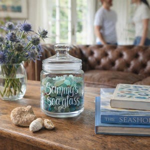 May include: A clear glass jar filled with blue and green sea glass, labelled "Sammy's sea glass" in white script. The jar is on a wooden table with a vase of blue flowers, seashells, and books.