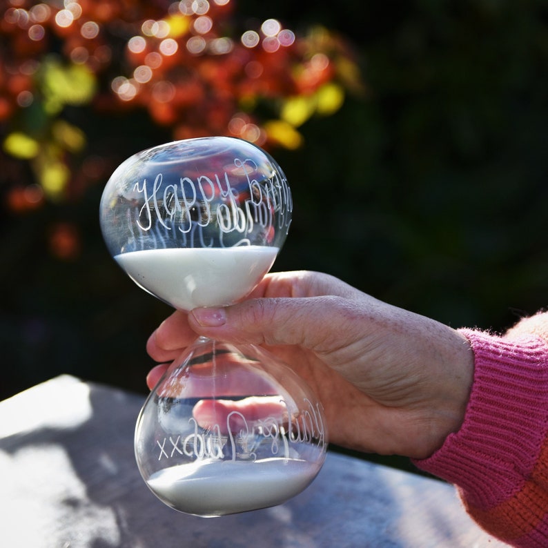 May include: Clear glass hourglass with white sand, held in a hand. The top of the hourglass reads "Happy Birthday" in cursive script. The bottom reads "XX Cool Grandma". The background is blurred with fall colors.