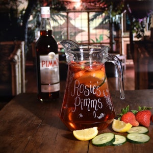 May include: A glass jug filled with a red drink, garnished with fruit and cucumber slices. The jug is etched with the words "Rosie's Pimms".