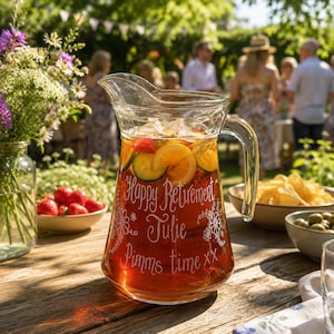 May include: A clear glass pitcher filled with amber liquid, ice, and slices of orange and cucumber. The glass is etched with white floral designs and the text: Happy Retirement, Julie, Pimms time xx. The pitcher has a sturdy handle and spout.