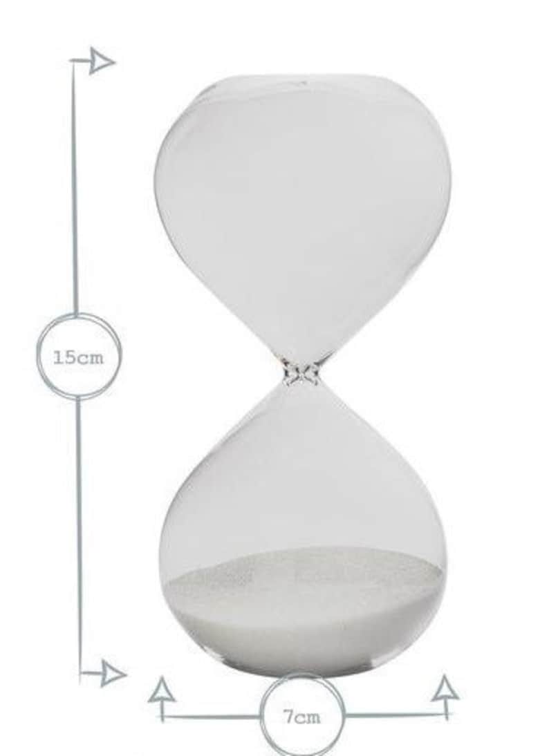 May include: Clear glass hourglass with white sand. The hourglass is 15cm tall and 7cm wide. The design features a classic shape with rounded top and bottom chambers connected by a narrow neck. The sand is visible inside the bottom chamber.