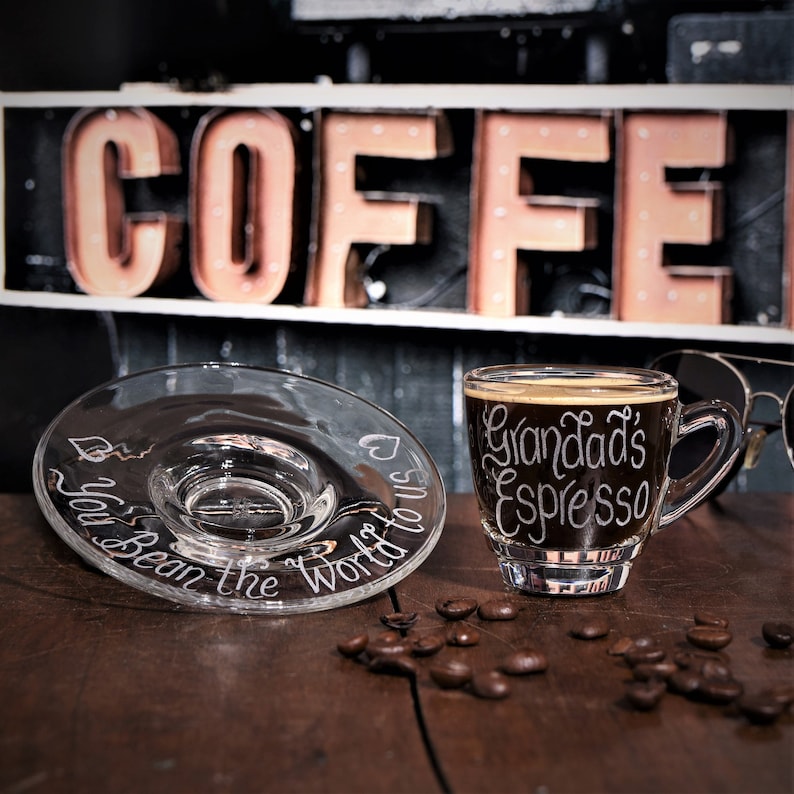 Glass espresso cup and saucer are shown in a coffee bar. The cup and saucer have both been hand engraved in a calligraphy font. The wording on the cup reads Grandads Espresso, and the saucer reads You bean the world to us with a heart design