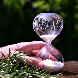 May include: Clear glass hourglass with white sand, inscribed with "50th Birthday" in elegant script. The hourglass is held by a hand, with green foliage in the foreground. The background is a blurred green.