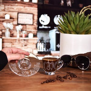 May include: A clear glass espresso cup and matching saucer set. The saucer is etched with the text Happy 30th Birthday in a cursive font. The espresso cup features an etched monogram with the letters CJM in a decorative script style.