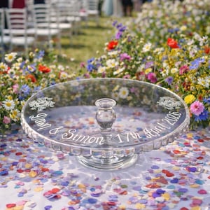 May include: A clear glass cake stand featuring a central pedestal base and a scalloped edge. The top surface is engraved with the text "Dawn & Simon 17th April 2026" in a cursive script, accompanied by two decorative floral motifs on opposite sides.