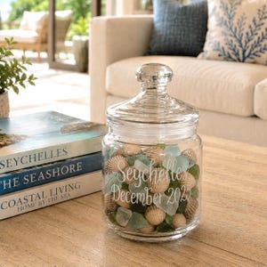 May include: A clear glass jar filled with seashells and sea glass, topped with a glass lid. The jar is inscribed with "Seychelles December 2025". The jar sits on a wooden table, with books visible in the background.