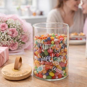 May include: A clear glass cylindrical jar filled with colourful assorted sweets. The front of the jar features white text reading Mummy's Sweet jar with sweet illustrations. A round wooden lid with a leather strap sits beside the jar.
