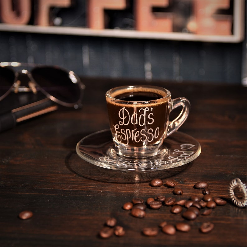 May include: A clear glass espresso cup with the words "Dad's Espresso" etched on the side. The cup is filled with dark coffee and sits on a clear glass saucer. Coffee beans are scattered on a dark wooden surface.