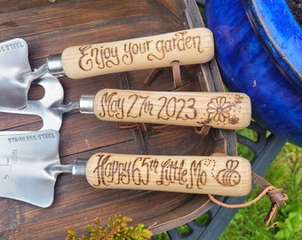 May include: Three stainless steel garden trowels with light wood handles featuring wood-burned text and designs. The handles are engraved with "Enjoy your garden," "May 27th 2023" with a flower, and "Happy 65th Little Mo" with a bee.