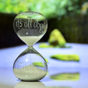 May include: A clear glass hourglass with white sand, featuring the etched text "it's all about" on top and "the timing" on the bottom. The hourglass is set against a blurred green and blue background.