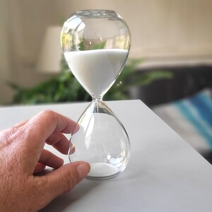 May include: Clear glass hourglass with white sand. The hourglass has a bulbous top and bottom, connected by a narrow neck. The sand is flowing from the top to the bottom. The hourglass is sitting on a gray surface.
