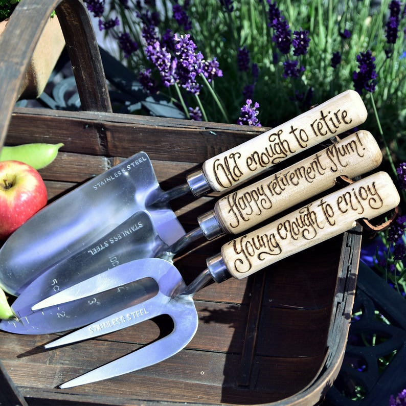 May include: A set of stainless steel garden tools with wooden handles. The handles are engraved with the phrases "Old enough to retire", "Happy retirement Mum!", and "Young enough to enjoy". The tools are in a wooden basket.