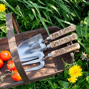 May include: A wooden basket filled with gardening tools and ripe tomatoes. The tools include a trowel, a hand fork, and a small shovel, all with wooden handles. The handles are engraved with phrases like "Thyme to Garden." The basket sits in a bed of green grass.