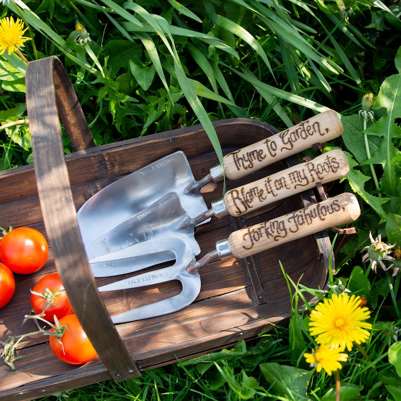 May include: A wooden basket filled with gardening tools and ripe tomatoes. The tools include a trowel, a hand fork, and a small shovel, all with wooden handles. The handles are engraved with phrases like "Thyme to Garden." The basket sits in a bed of green grass.