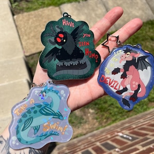 May include: Three acrylic keychains featuring illustrated creatures. One is a black moth-like figure with red eyes and the text HAVE YOU SEEN HIM?. Another is a pink horned creature with wings and the text LET IT BE THE DEVIL!. The third is a blue sea serpent with the text Take A Swim!.