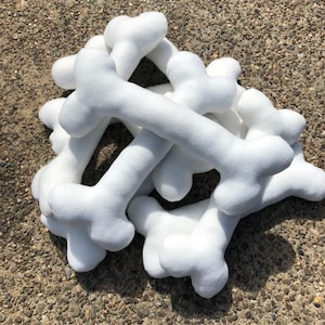 May include: A pile of white plush dog bones. The bones are soft and fluffy, with a classic bone shape. They are scattered on a textured surface, suggesting an outdoor setting. The bones are a solid white color.