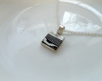 Op de afbeelding: Een zilveren ketting met een vierkante hanger met een zwart-wit gestreept patroon.