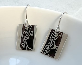 Op de afbeelding: Een paar zilveren oorhangers met een rechthoekige hanger in zwart en zilver. De hanger heeft een golvend, abstract ontwerp.