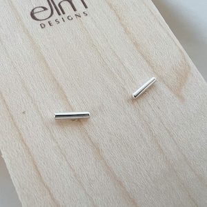 May include: A pair of silver bar stud earrings on a wooden surface with the text "elm designs" on the surface.