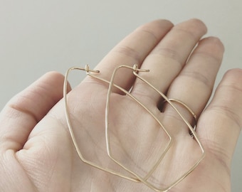 Puede incluir: Un par de pendientes de aro geométricos dorados. Los pendientes tienen una forma angular única, con una textura martillada. Los aros se sostienen en una mano, mostrando su diseño y artesanía. Los pendientes son un accesorio elegante.