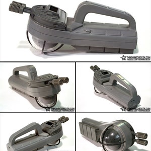 May include: A gray prop weapon with a handle, two cylindrical barrels, and a clear, rounded front. The design includes a top section with a rectangular window and a handle for gripping. The text "THROWINGCHICKEN.COM" is visible.