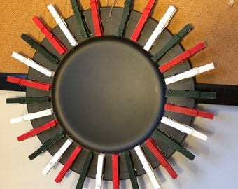 Puede incluir: Un plato circular de metal negro con una disposición en forma de sol de pinzas de madera rojas, blancas y verde oscuro alternadas, sujetas alrededor de todo el borde exterior. Incluye un trozo de cuerda en la parte superior para colgar.