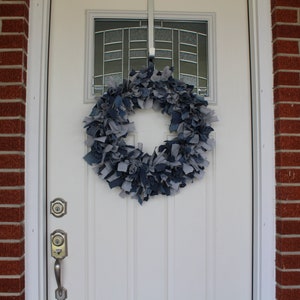 May include: A blue and gray denim wreath hangs on a white front door with a glass window pane. The wreath is made of strips of denim fabric.