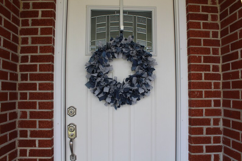 May include: A blue and gray denim wreath hangs on a white front door with a glass window pane. The wreath is made of strips of denim fabric.