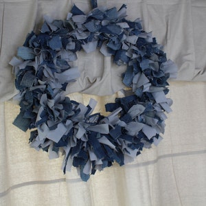 May include: A blue and gray denim wreath made from strips of fabric. The wreath is hanging on a white wall.