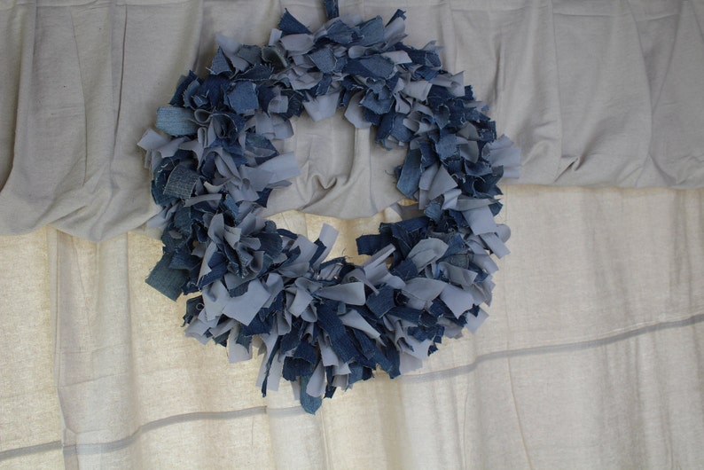 May include: A blue and gray denim wreath made from strips of fabric. The wreath is hanging on a white wall.