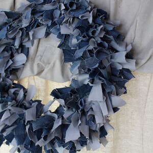 May include: A wreath made from strips of blue and gray denim fabric. The wreath is hanging on a white wall.