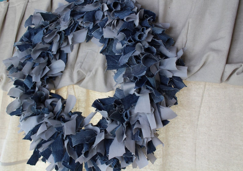 May include: A wreath made from strips of blue and gray denim fabric. The wreath is hanging on a white wall.