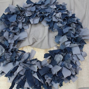 May include: A blue and gray denim wreath made from strips of fabric. The wreath is hanging on a white wall.