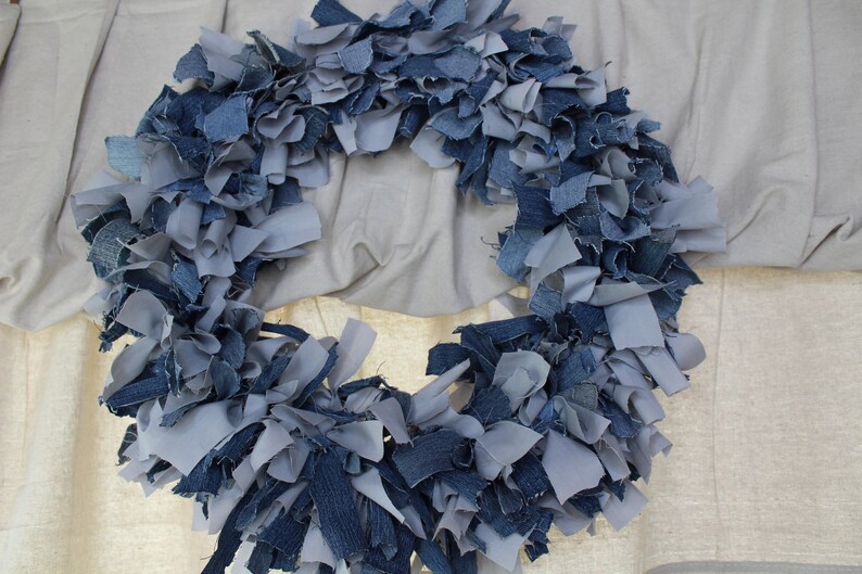 May include: A blue and gray denim wreath made from strips of fabric. The wreath is hanging on a white wall.